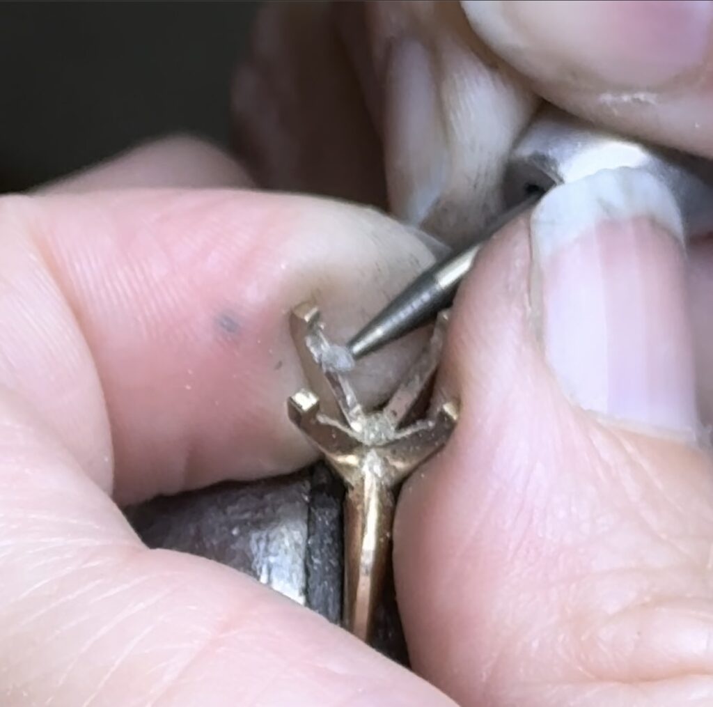 A close-up of a jeweler's hand working on a ring with a fine tool, showcasing intricate design details.