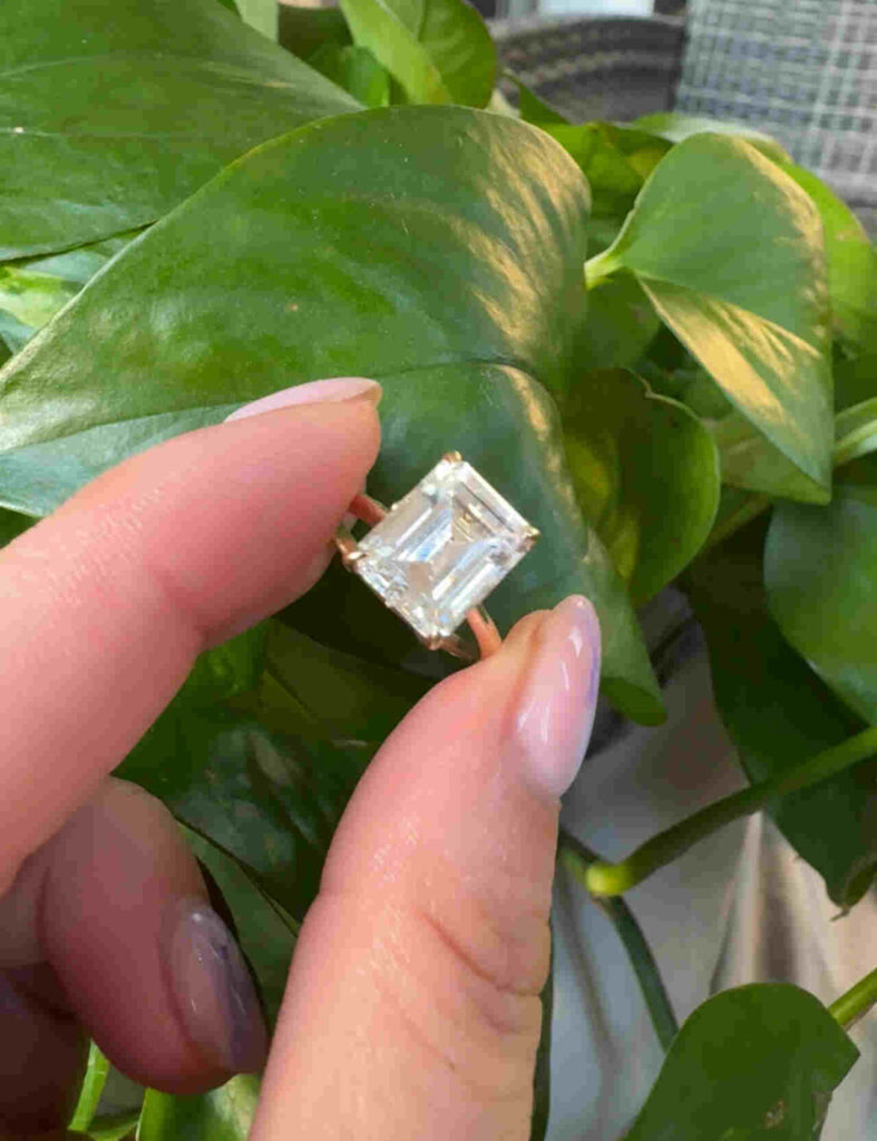 Emerald cut classic ring holding in hand in front of green leave, display classic and clear diamond by gaia jewelry