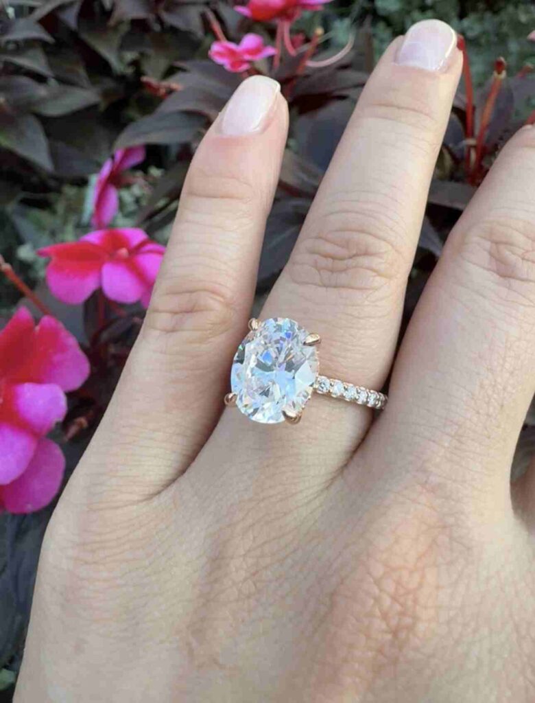 Oval shaped diamond ring by gaia jewelers displayed in a finger with a pink flowers in background.